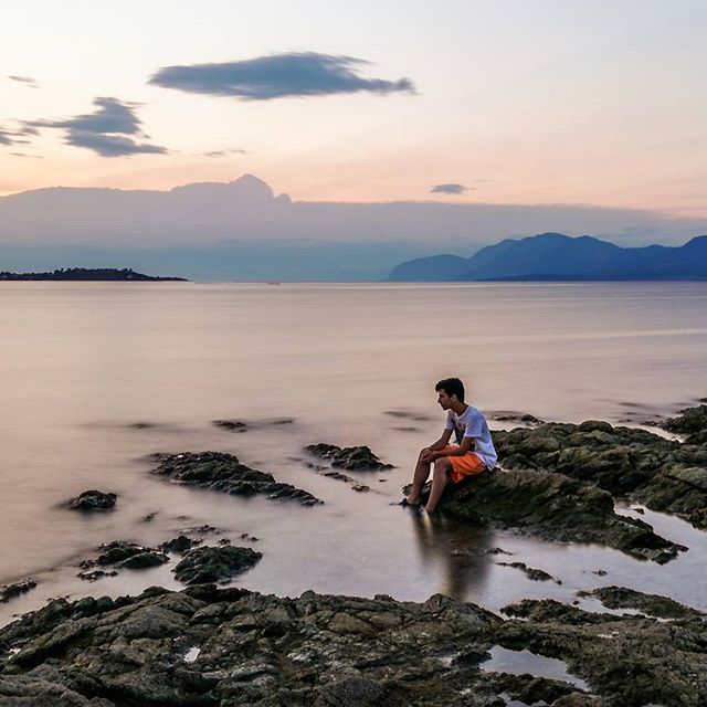 A person sits on the rocky coast during a serene sunset over the sea and distant mountains.