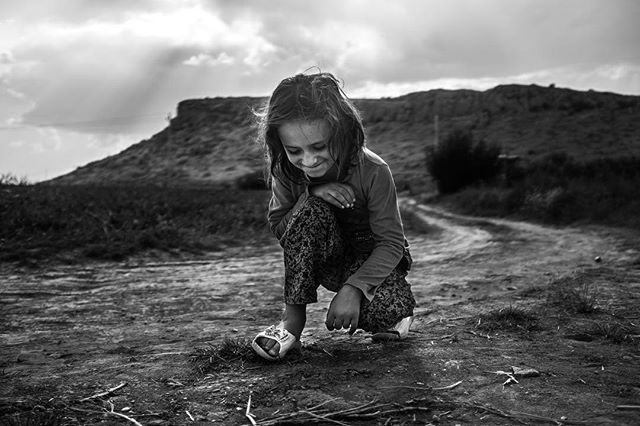 A young girl crouches on a dirt road, lost in thought, in a black and white image.