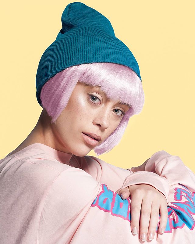 A woman with pink hair and a blue beanie poses in a studio against a yellow background.