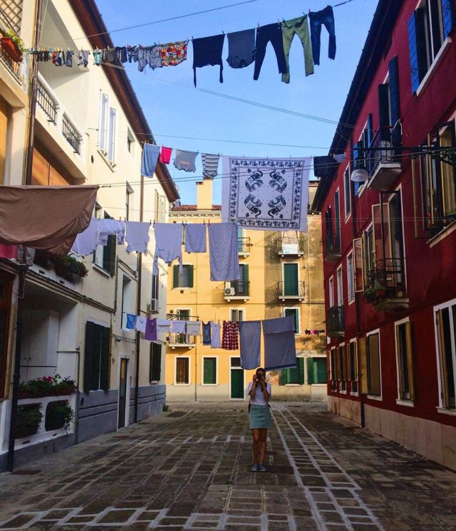 A woman stands in a European alleyway with laundry hanging overhead, capturing a glimpse of everyday life.