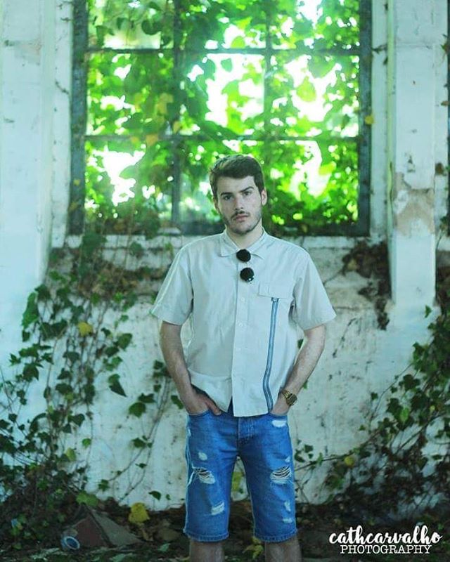 A man poses in an abandoned building in blue shorts near a window with vines.