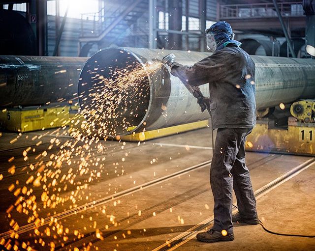 A metal worker welds a large pipe in an industrial setting with bright sparks flying around him.
