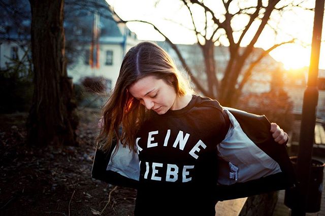 A brunette woman in a t-shirt poses outdoors in soft sunlight.
