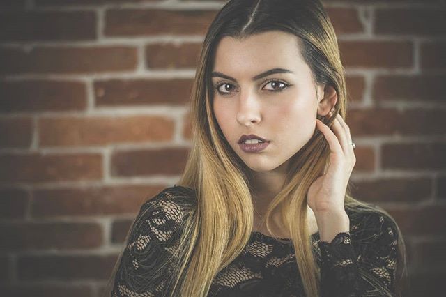 A beautiful woman poses in a studio against a brick wall with soft, diffused lighting and a shallow depth of field.