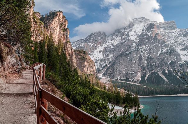 A scenic path with a wooden fence leads to a beautiful lake surrounded by majestic mountains and lush trees in Italy.