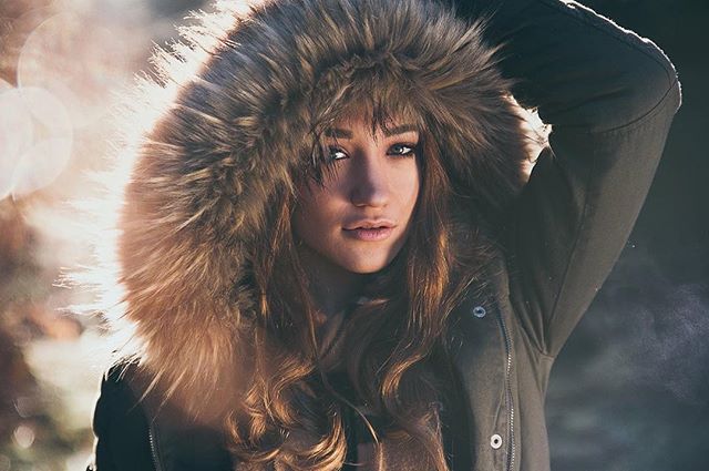 A woman in a fur-trimmed hood poses in soft, diffused light for a fashion editorial.