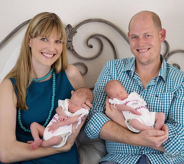 A smiling family of four, including newborn twins, poses indoors with soft, natural lighting.