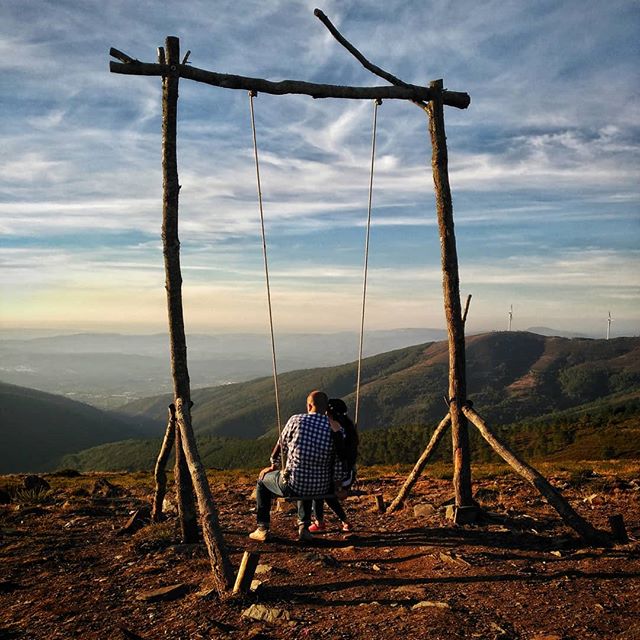 A couple enjoys a scenic view from a wooden swing atop a mountain, creating a peaceful and romantic landscape.