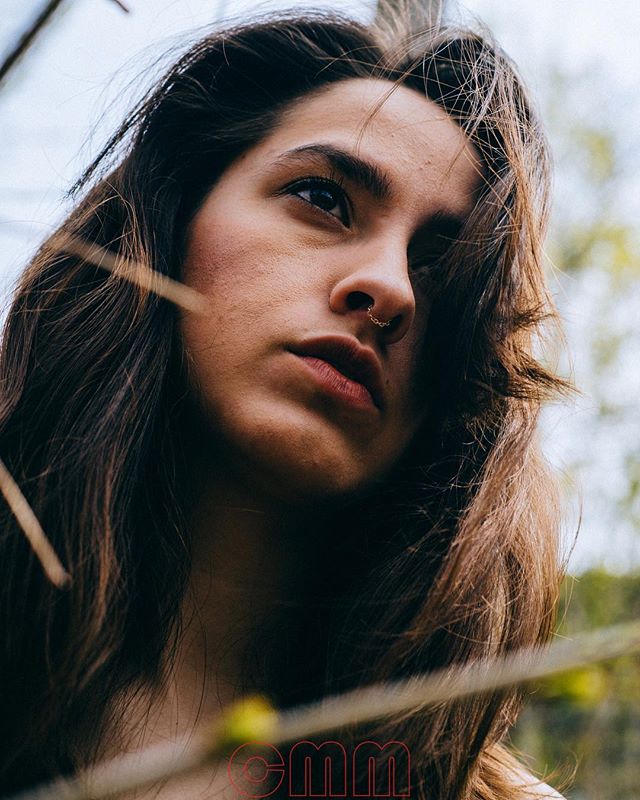 A brunette woman with a nose ring gazes off-camera with a contemplative expression amidst a natural outdoor setting.