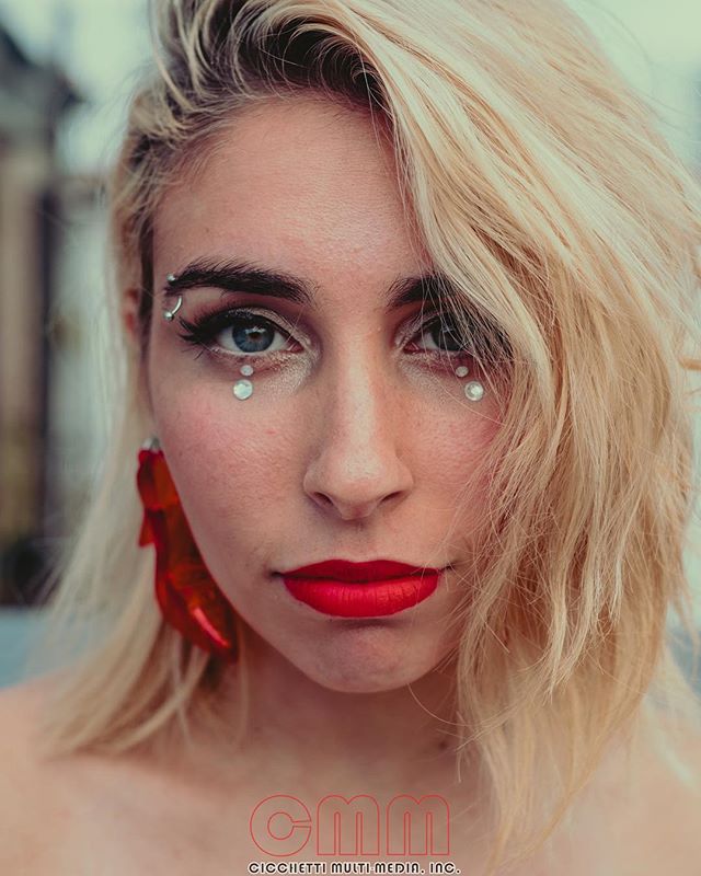 A woman with blonde hair, red lipstick, and face gems stares intensely at the camera.