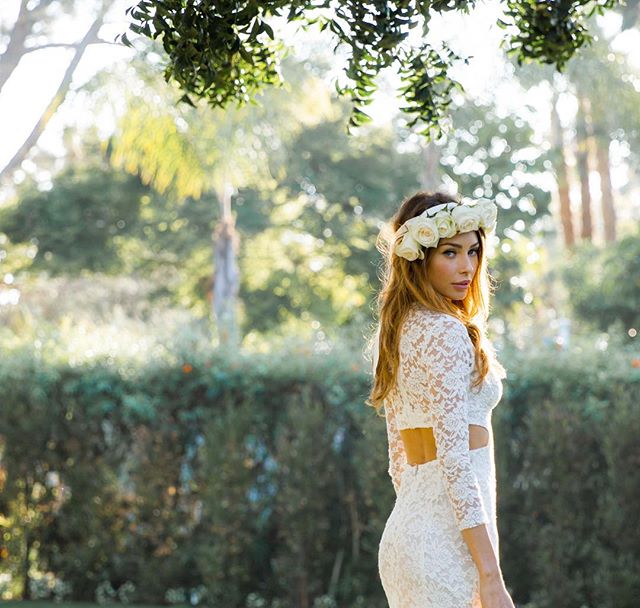 A woman in a white lace dress and flower crown poses in a lush green garden.