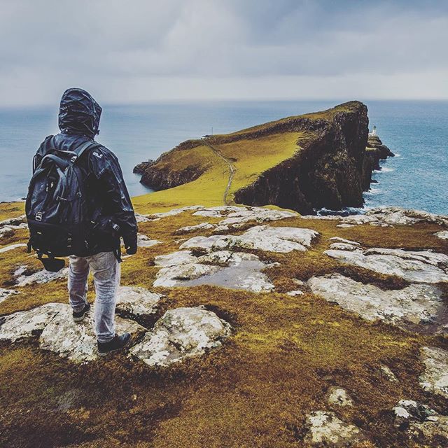 A lone hiker stands on a rocky cliff overlooking the ocean in a scenic, moody landscape.