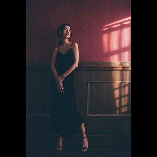 A woman in a black dress stands in a studio with light casting shadows on the wall.