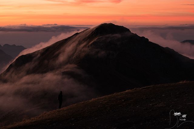 A lone figure stands on a mountain ridge during a serene sunrise, enveloped by soft clouds.