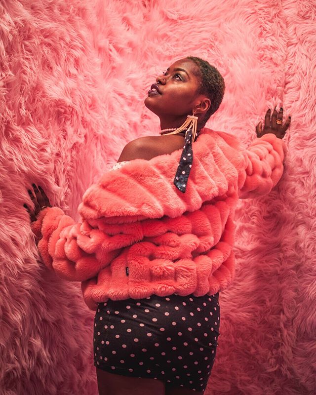 A stylish woman poses against a pink backdrop in a faux fur coat and polka dot skirt.
