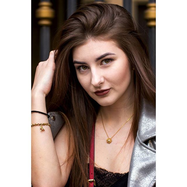 A stylish brunette woman poses outdoors wearing a necklace and a silver jacket.
