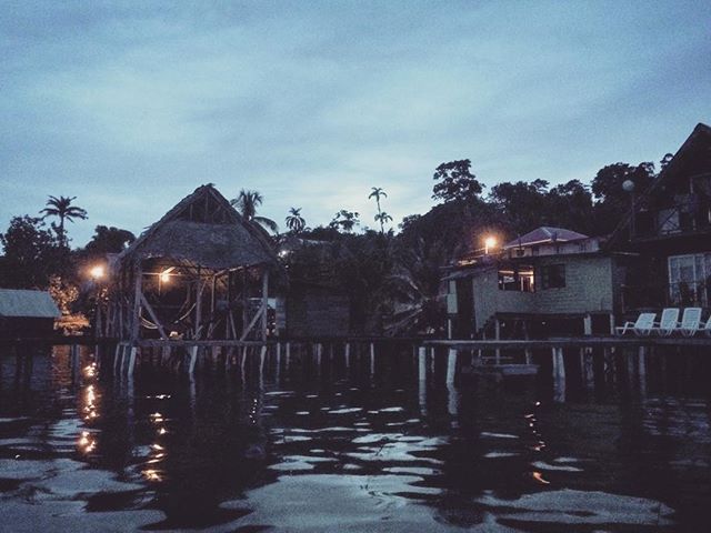 Tropical houses on stilts reflect in tranquil water, under a dusky sky, creating a serene waterfront scene.