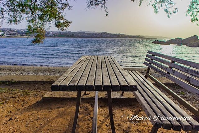 A serene seascape features a wooden table and bench overlooking the calm ocean waters and distant coastline.
