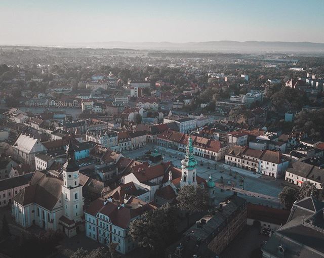 An aerial view captures a European town with red-roofed buildings, a central plaza, and distant hills under a hazy sky.