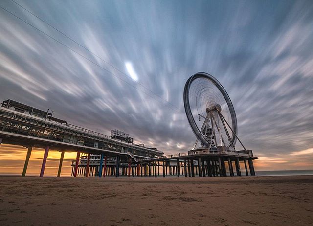 Scenic view of a Ferris wheel and pier on a sandy beach, captured with long exposure under a cloudy sky.
