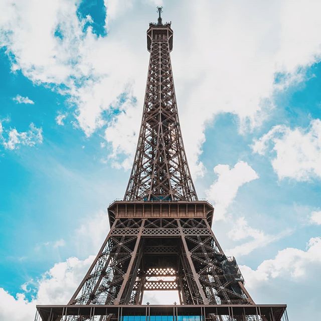 A low angle view of the Eiffel Tower against a bright, partly cloudy blue sky in Paris, France.