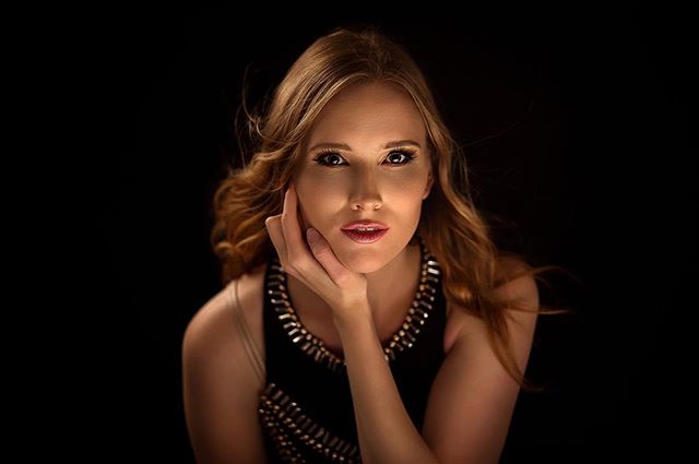 Elegant woman with blonde hair poses against a dark background, her hand gently touching her face.