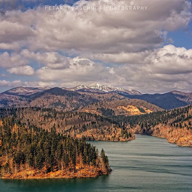 Scenic view of a lake surrounded by a lush forest and majestic mountains under a partly cloudy sky.