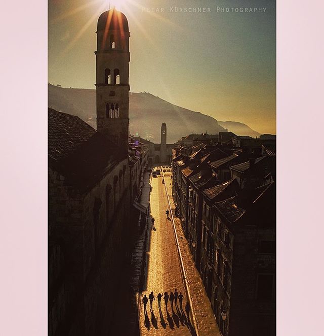 A long view down a sunlit European street with people walking towards a historic tower and old buildings.