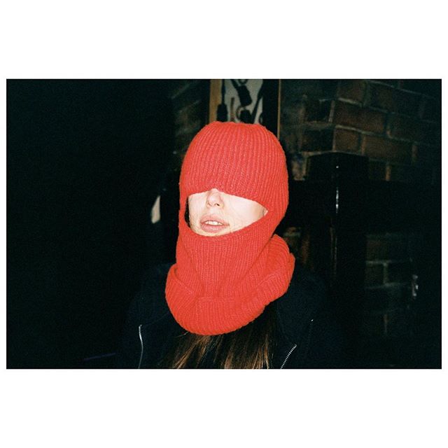 A person wearing a red knitted balaclava stands in a dimly lit indoor setting.