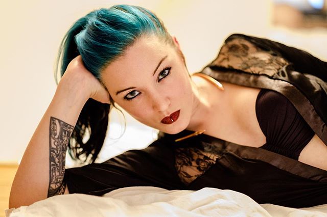 A woman with blue hair and a tattoo poses in a sensual portrait with soft, warm lighting indoors.