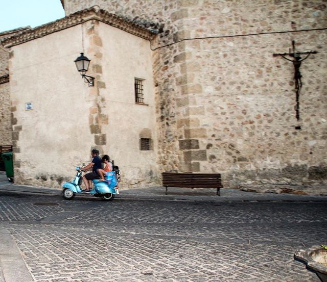 A man and woman ride a blue scooter through a historic European city on a cobblestone street by an old stone building.