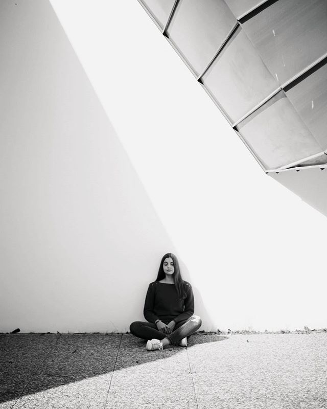 A woman sits peacefully against a wall in a monochrome setting, bathed in soft, natural light.