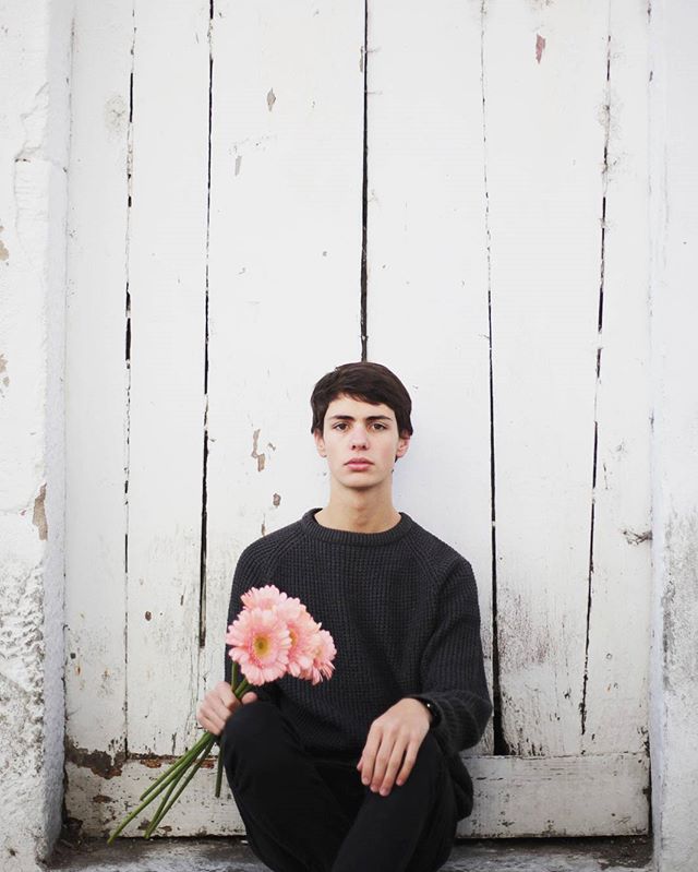 A young man in dark clothing sits holding pink gerbera daisies against a white weathered wall.