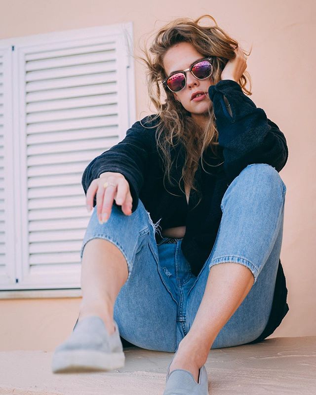 A fashionable woman sits confidently wearing sunglasses, a jacket, and jeans for an urban, relaxed style.