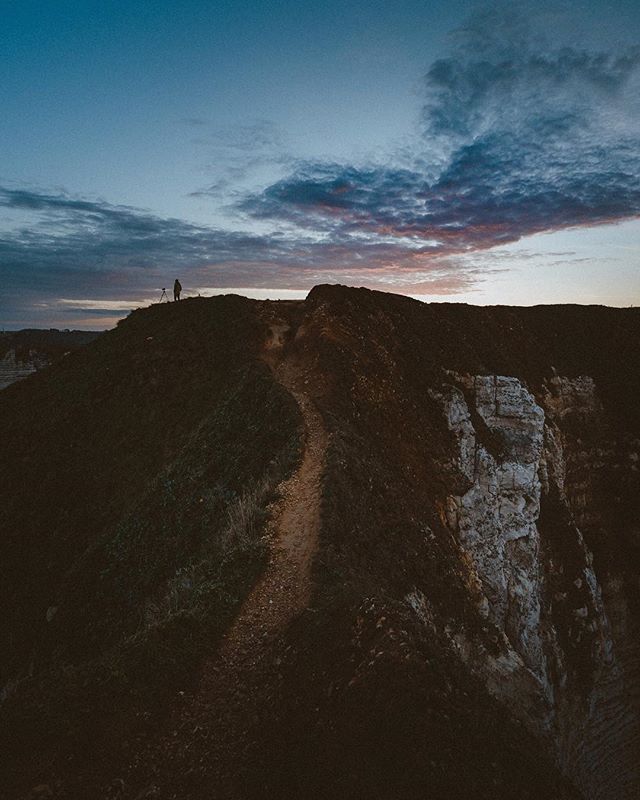 A lone person sets up a tripod on a hilltop path at dusk with a scenic cliffside view.