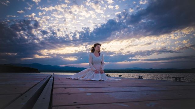 A woman sits on a pier in a dress, enjoying the peaceful lake and mountain view under a cloudy sky.