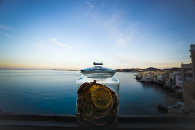 A glass jar filled with dried citrus fruit sits against a scenic backdrop of the sea and a distant cityscape.