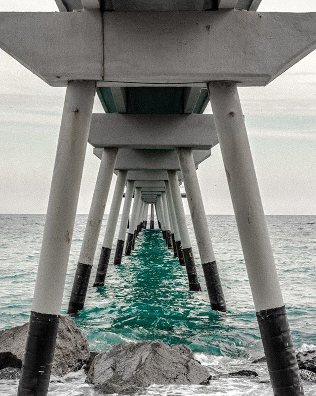 View through the supports of a pier with calming teal ocean water, captured with a low angle.