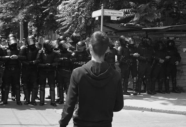 A man stands facing a line of riot police during a protest in a black and white image.