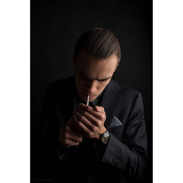 A man in a suit dramatically lights a cigarette against a dark backdrop, evoking a sense of mystery and sophistication.