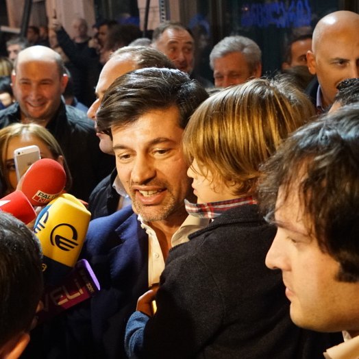 A man and child are surrounded by reporters with microphones in an urban setting, captured in a candid, news style photograph.