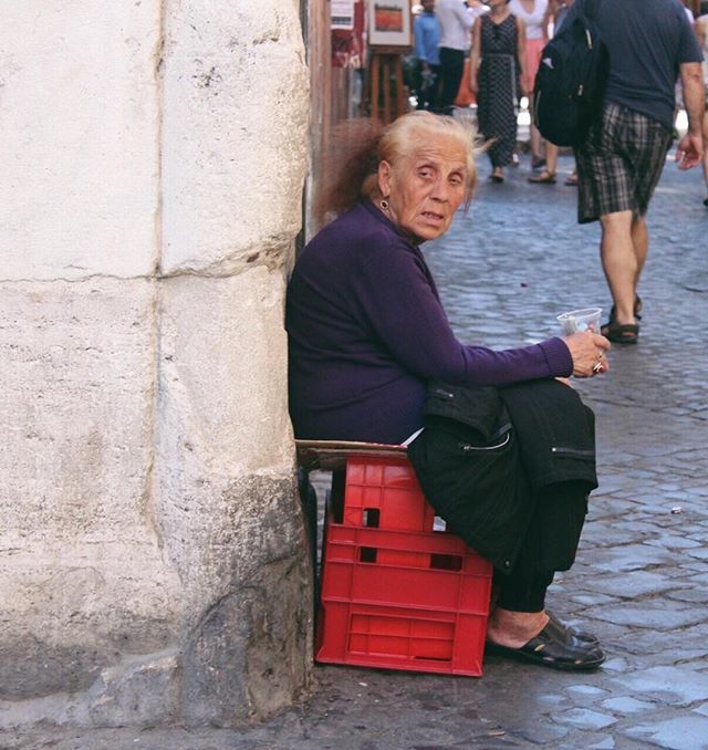 An elderly woman sits on crates on a city street looking thoughtful.