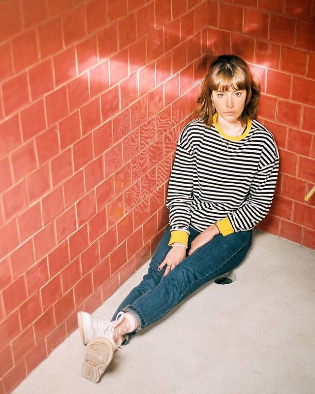 A woman in a striped shirt and jeans sits in a red-tiled corner, looking serious and stylish.