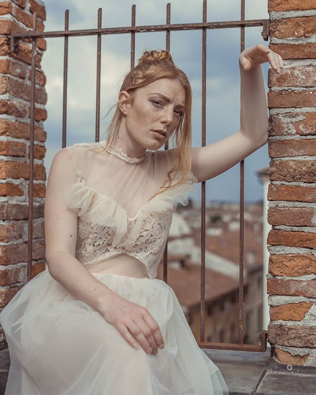 A redhead woman in a lace dress poses on a rooftop with a cityscape in the background.