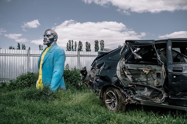 A Lenin statue stands beside a wrecked car in a field, symbolizing political and societal upheaval.