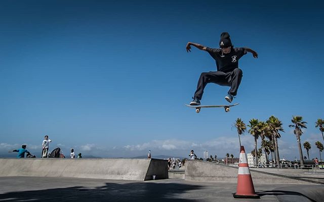 A skateboarder performs a jump at a skatepark under a clear blue sky, capturing an energetic urban scene.