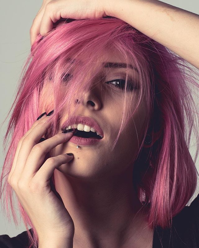 A woman with pink hair poses with hand near her mouth, showcasing a confident and edgy style.