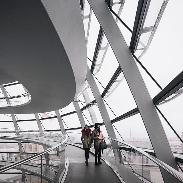 Two people walk inside a modern architectural dome with glass and steel on a cloudy day.