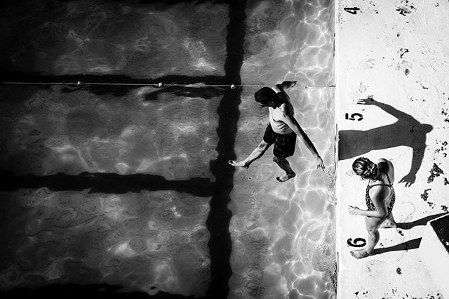 A black and white high-angle shot shows two people near a swimming pool, one about to dive.