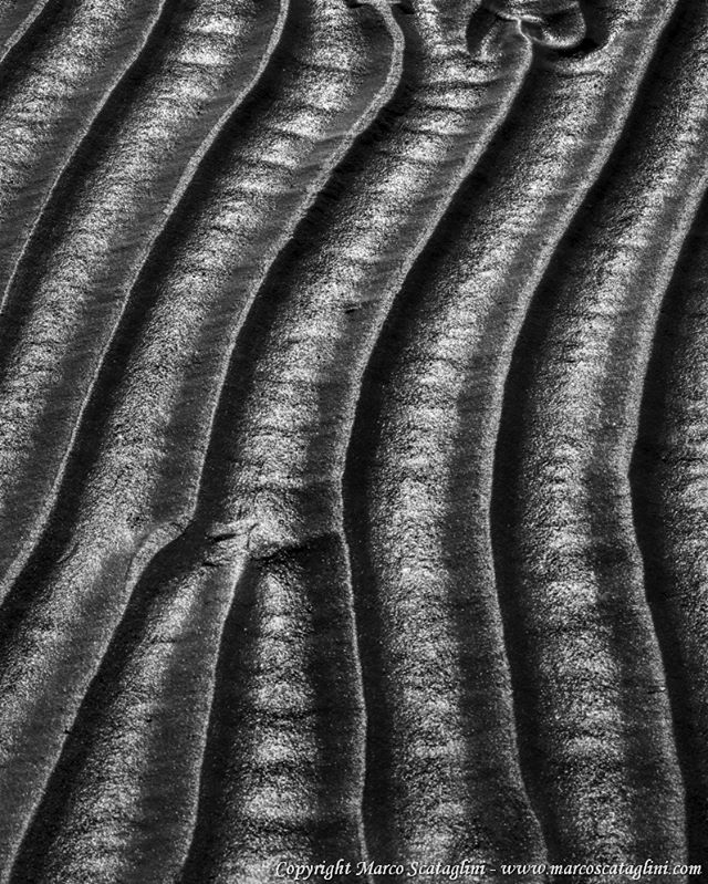 Black and white close-up of wave patterns in the sand at the beach, creating an abstract textured background.